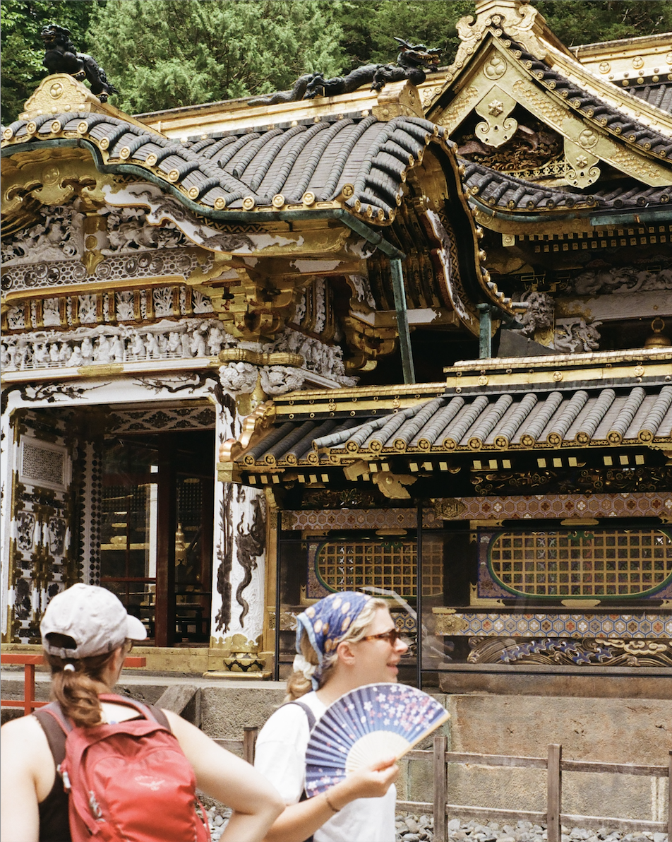 Two tourists in front of an ornate traditional Japanese temple with intricate golden and black roof details.