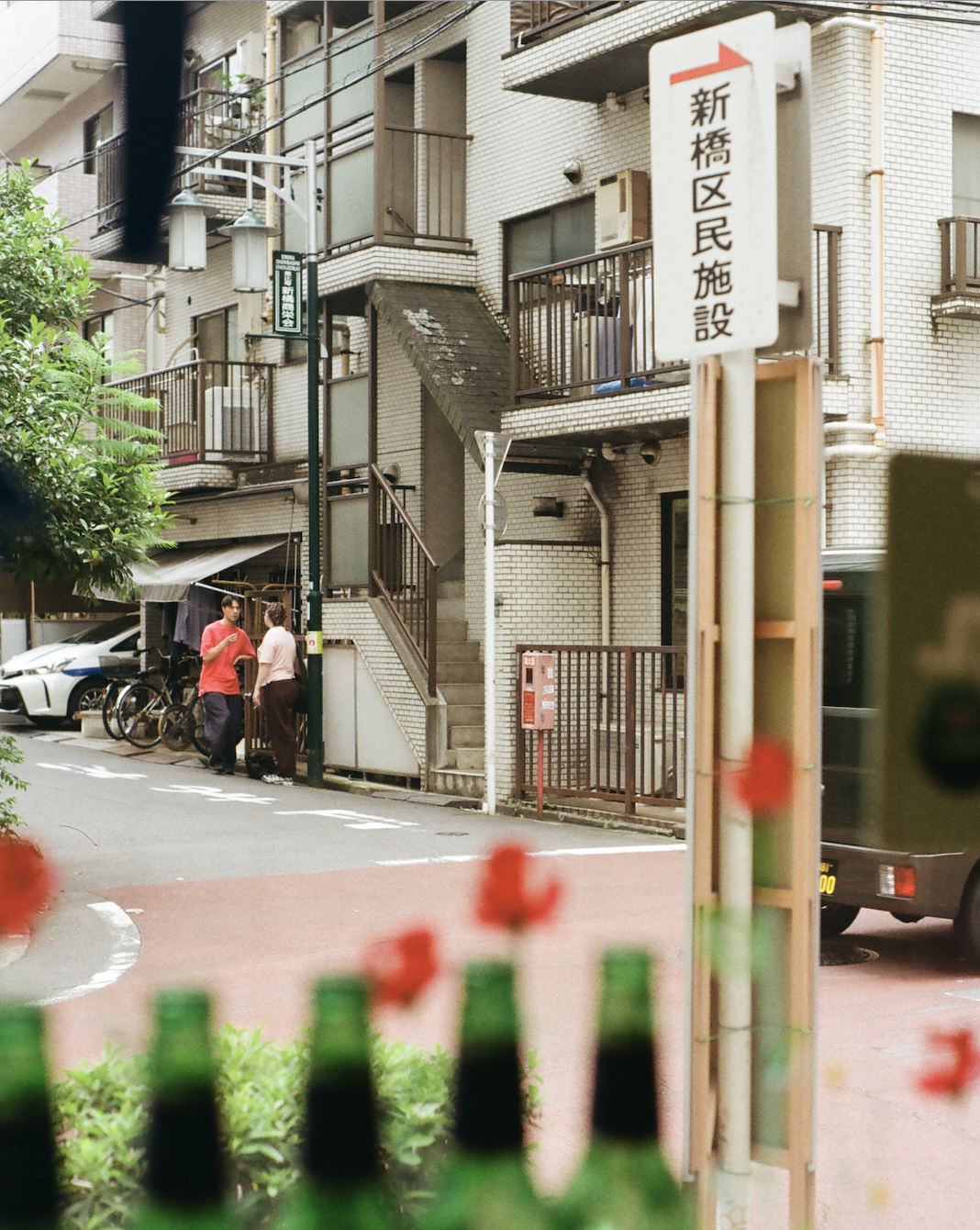 Two people talking on a quiet street corner near a white brick residential building with balconies and staircases, with green bottles and red flowers blurred in the foreground.