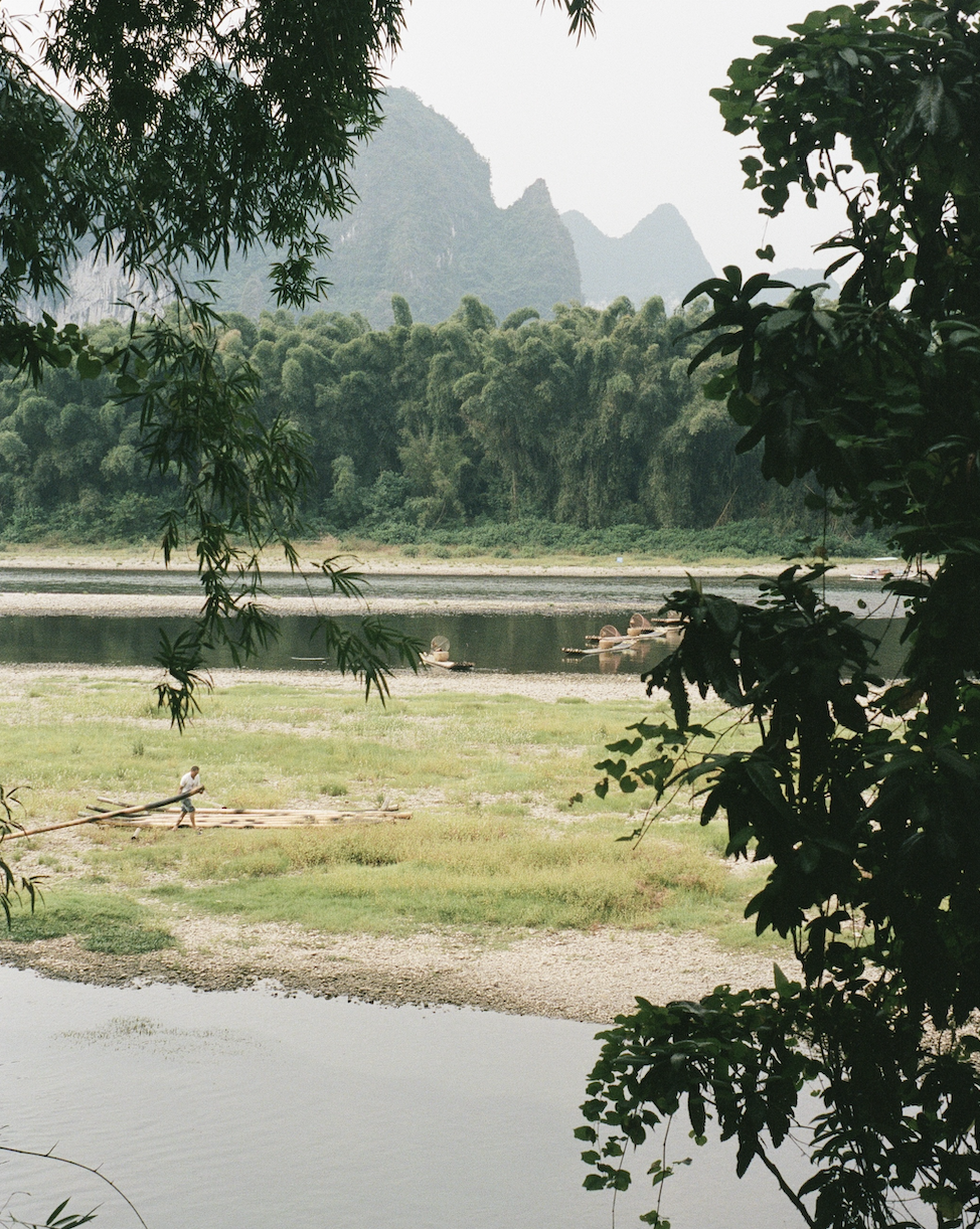 A person standing on a bamboo raft near a riverbank with lush green trees and forested hills in the background.