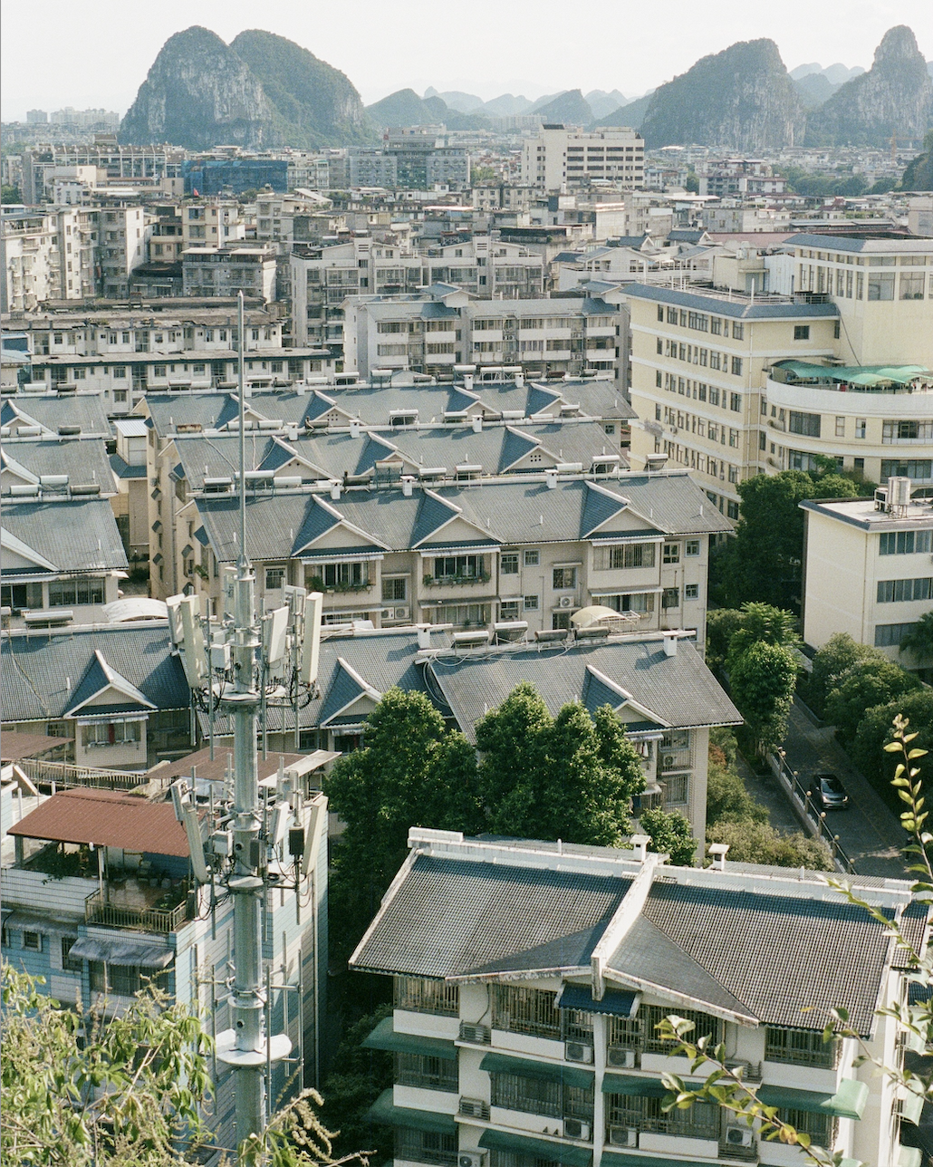 Residential buildings with traditional gray-tiled roofs in the foreground and green trees, with karst mountains and more city buildings in the background.