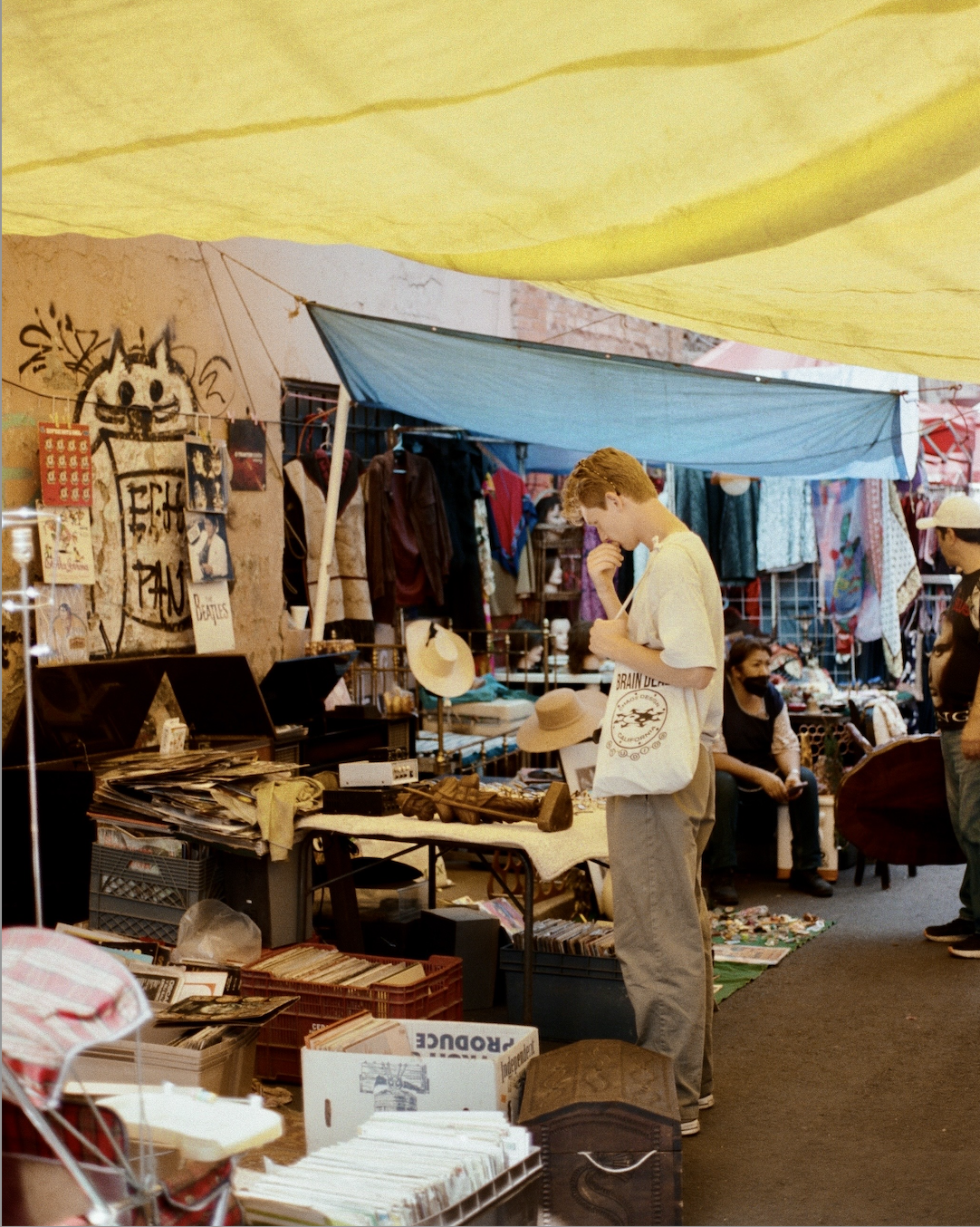 A young man with a tote bag browsing records at an outdoor market stall with various vintage items and clothing around.