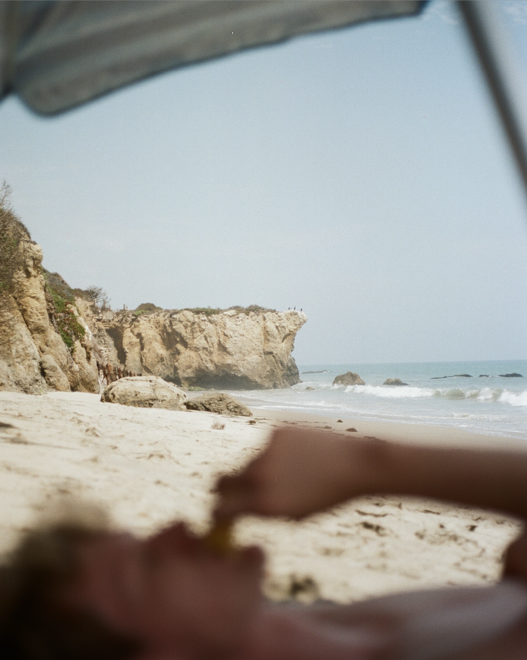 Sandy beach with rocky cliffs in the background and a blurred person lying under an umbrella in the foreground.