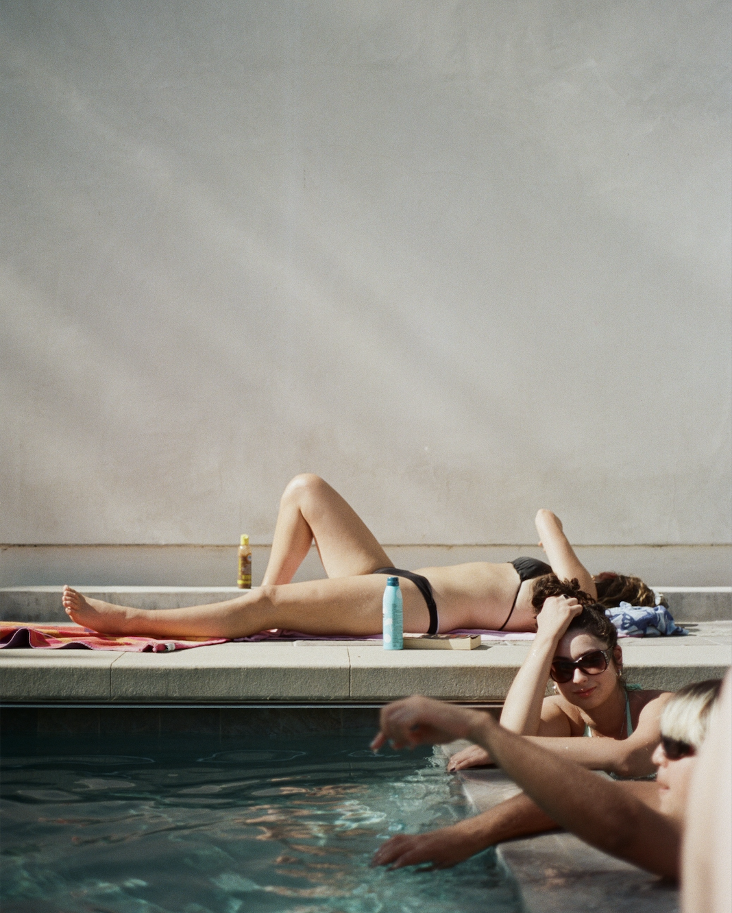 Three people relaxing by a pool; one woman in a black bikini sunbathes on a towel, while two others lean on the pool edge, one wearing sunglasses.
