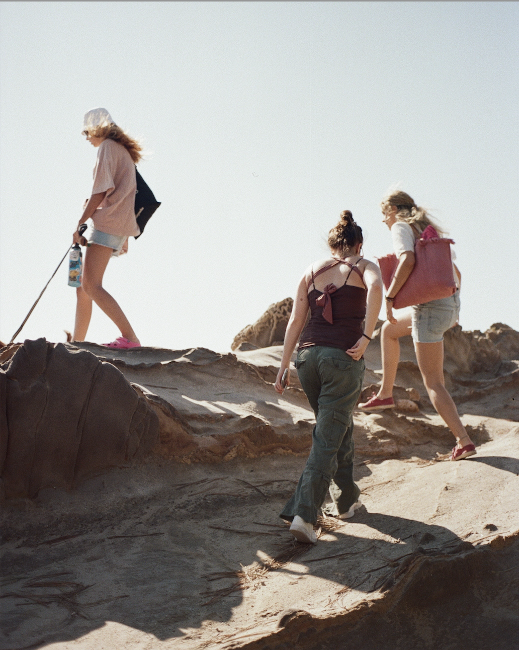 Three women hiking up rocky terrain under clear sky, one holding a water bottle and walking stick.