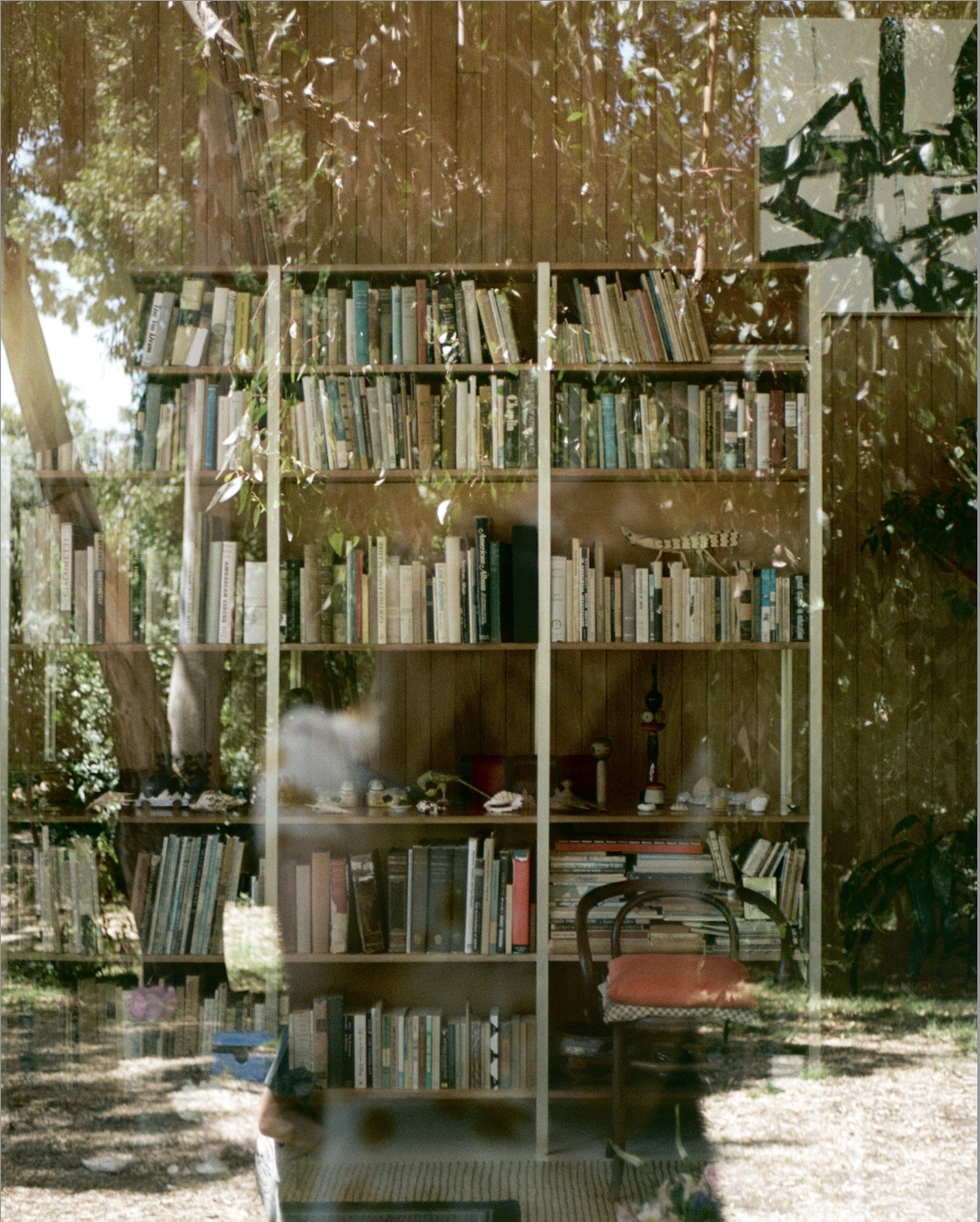 Bookshelf filled with books and decorative items reflected through a window showing outdoor greenery and a wooden chair with an orange cushion.