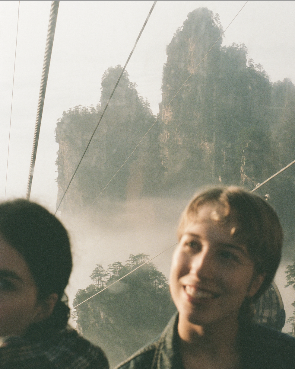 Two people on a cable car with misty, forested mountainous peaks in the background.