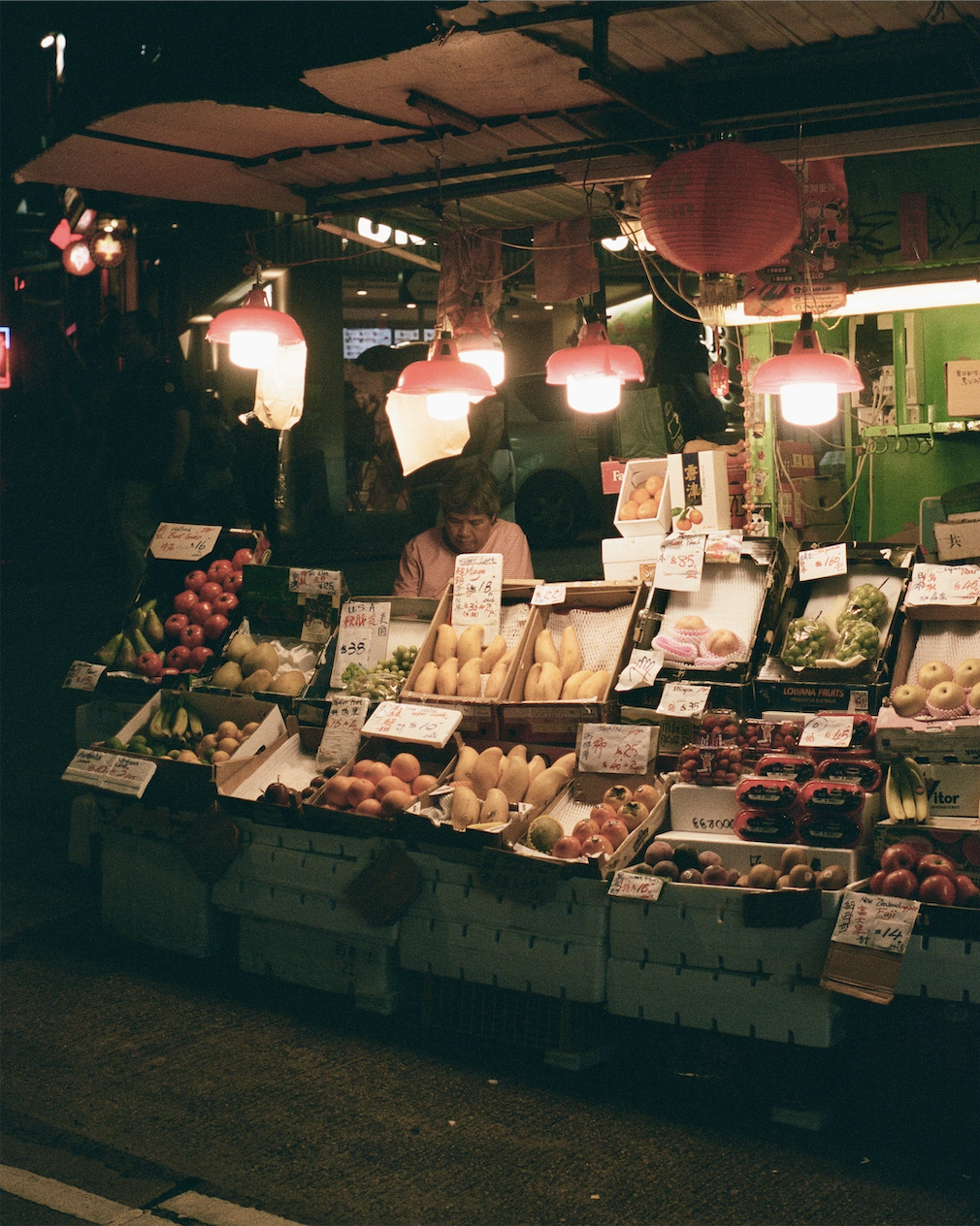 Night market stall brightly lit with red hanging lamps displaying various fruits and a vendor sitting behind the counter.