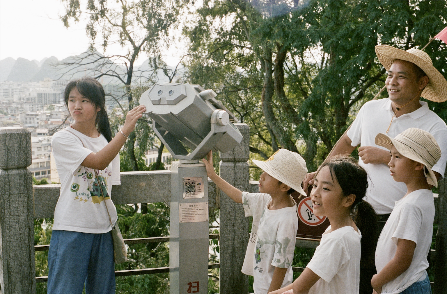 Group of children and a man wearing straw hats standing outdoors, with two kids interacting with a viewing telescope on a scenic overlook.