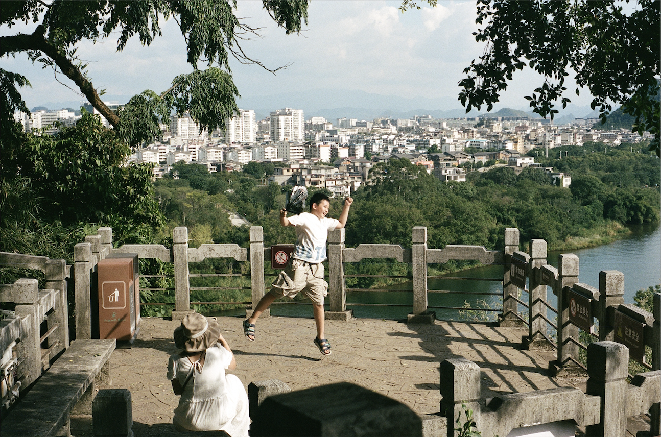 Child joyfully jumping in the air on a stone platform with a city and trees in the background, while a person in a hat sits nearby.