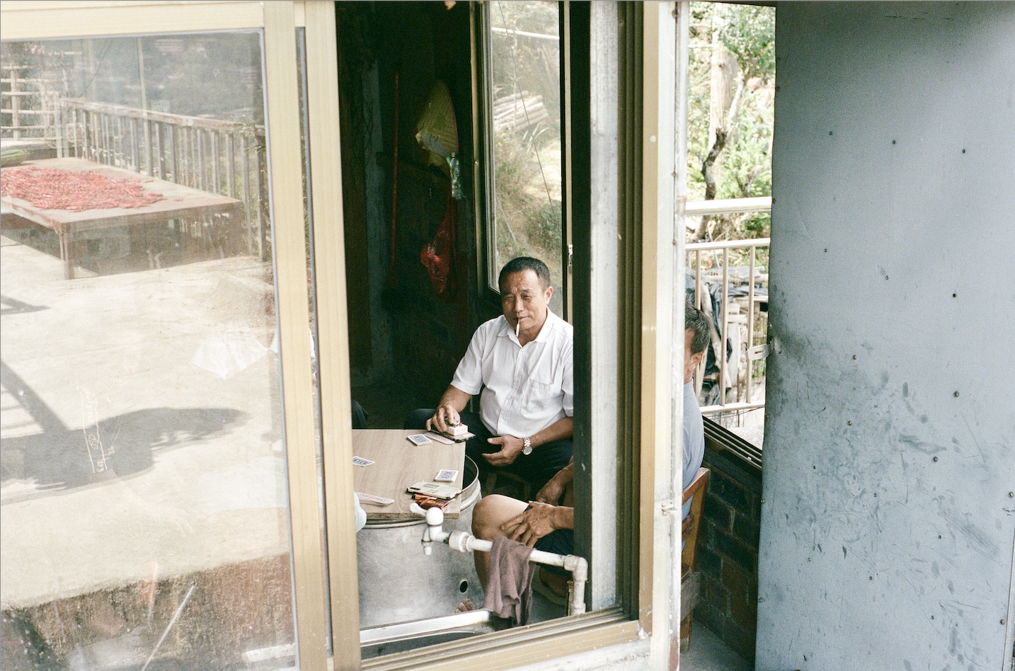Two men sitting inside a small room playing cards around a round table, with one man smoking a cigarette.