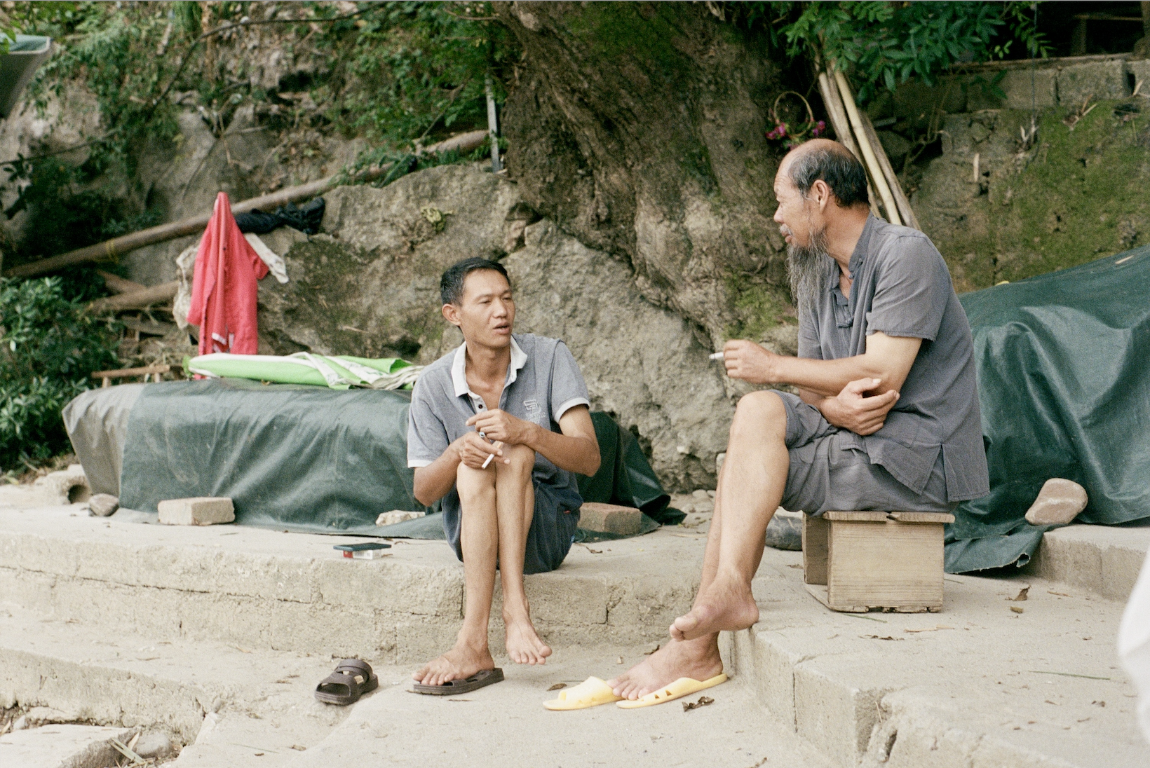 Two barefoot men sitting outdoors on stone steps, smoking and talking near a rock and greenery.