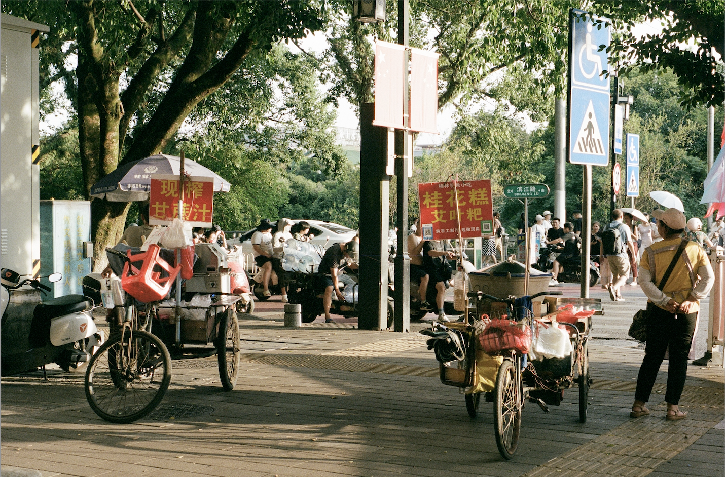 Street scene with two tricycles carrying goods and red signs in Chinese, people sitting and walking under trees near crosswalk and disability signs.